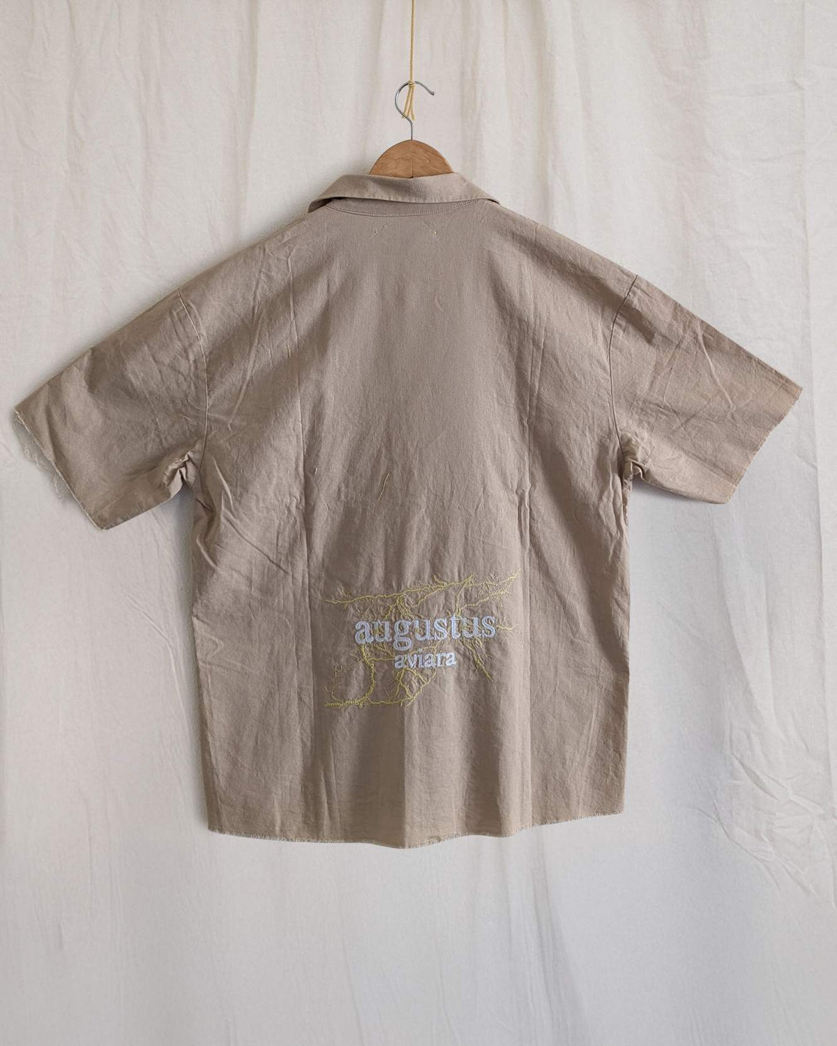 Beige shirt with 'Augustus' branding on a hanger against a white background